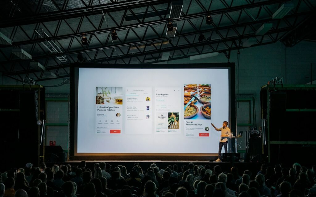 Person discussing while standing in front of a large screen in front of people inside dim-lighted room